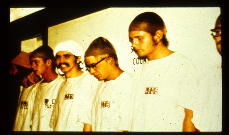 A grainy, candid black and white historical photograph from the 1971 Stanford Prison Experiment. The scene captures a narrow, dimly lit basement hallway converted into a makeshift 'prison.' Several young men, identified as prisoners by their white gowns and numbers, are huddled together on a low cot inside one of the 'cells,' their heads bowed. A guard, wearing a dark khaki uniform, a hat, and reflective aviator sunglasses, stands authoritatively in the foreground corridor over them, holding a wooden nightstick and pointing. The walls are concrete block with hand-painted numbers and bars on small window openings.